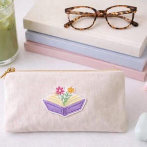 May include: A light beige pencil case with a gold zipper, featuring an embroidered open book design with flowers. Tortoiseshell glasses rest on a stack of books. A glass of green drink, a hair clip, and a case are also visible.