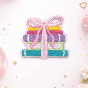 May include: Embroidered patch featuring a stack of colorful books tied with a pink bow. The books are in shades of yellow, pink, purple, green, and blue. White stitching outlines the design, creating a charming, decorative accent.