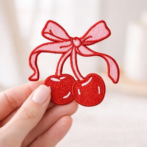 May include: Embroidered patch featuring two red cherries with white highlights and a pink bow. The patch is a decorative item, suitable for clothing or accessories. The design is a playful, whimsical representation of fruit.