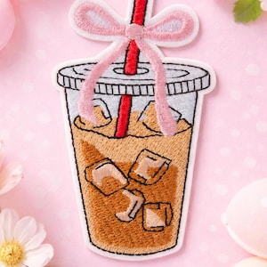 May include: Embroidered patch of an iced coffee drink in a clear plastic cup with a red straw and a pink bow. The coffee is light brown with ice cubes. The patch has a white border.