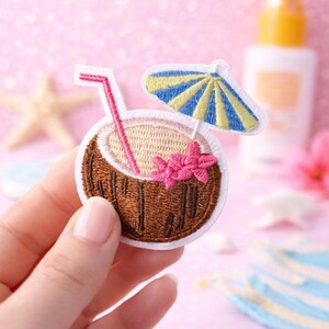 May include: Embroidered patch featuring a coconut cocktail with a pink straw, a blue and yellow umbrella, and pink flowers. The patch is brown, white, and pink, with a white border. The patch is held in a hand against a pink background.