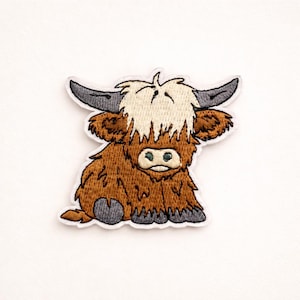 May include: Embroidered patch featuring a cartoon Highland cow. The patch has a white border and depicts the cow in shades of brown, gray, and cream. The cow is sitting with its legs tucked under its body, and has large horns.