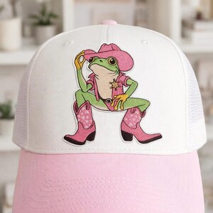 May include: White and pink trucker hat featuring an embroidered cartoon frog dressed as a cowboy. The frog wears a pink cowboy hat, shirt, and boots with heart patterns. The hat has a pink brim and a white mesh back.