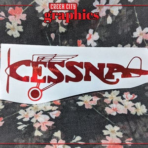 May include: Red vinyl decal of a Cessna airplane with the word "CESSNA" in bold letters. The decal is on a white background.