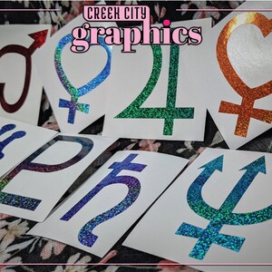 May include: A collection of holographic vinyl stickers featuring various symbols and designs, including hearts, planets, and astrological signs. The stickers are on a white background and have a glittery finish. The text "Creek City Graphics" is visible at the top of the image.