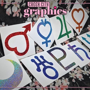 May include: A set of nine glitter stickers featuring various astrological symbols, including a crescent moon, a heart, a trident, and a cross. The stickers are on white paper and have a sparkly, iridescent finish.