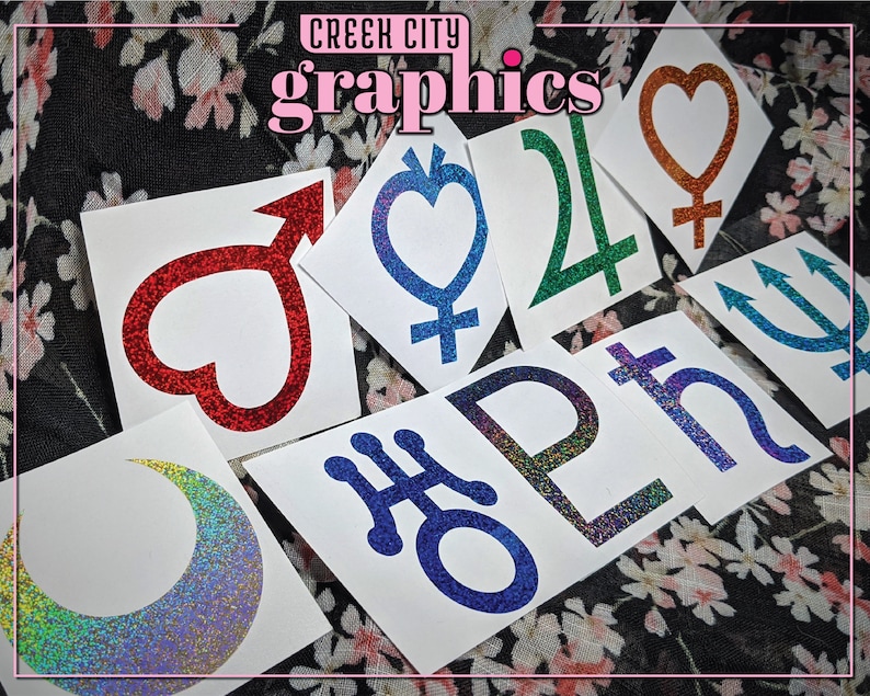May include: A set of nine glitter stickers featuring various astrological symbols, including a crescent moon, a heart, a trident, and a cross. The stickers are on white paper and have a sparkly, iridescent finish.