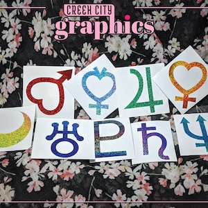 May include: A set of nine iridescent glitter stickers featuring various symbols, including a crescent moon, a heart, a trident, and a cross. The stickers are on a white background with a floral pattern.