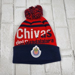 May include: A red and navy blue knit beanie with the text "Chivas del Guadalajara" in white. The beanie features a red pom-pom and a team logo patch.