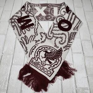 May include: A white and burgundy knitted scarf with the word "MEXICO" and a national emblem. The scarf has burgundy tassels and the word "M" and "O" on either end. The scarf is made of a soft, knitted material.