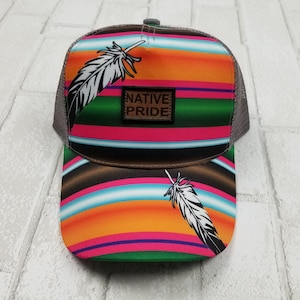 May include: A gray and brown trucker hat with a colorful striped pattern and a white feather design. The hat has a patch that says "NATIVE PRIDE" on it.