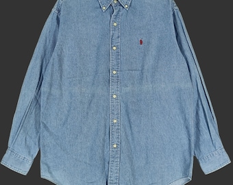 May include: Light blue denim button-down shirt with long sleeves. Features a button-down collar, a small red logo on the chest, and buttoned cuffs. The shirt has a curved hemline and a classic fit.