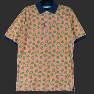 Op de afbeelding: Oranje poloshirt met een herhaald patroon van groene en paarse bloemmotieven. De kraag en mouwen zijn marineblauw. Het shirt is gemaakt van een zacht, comfortabel materiaal.