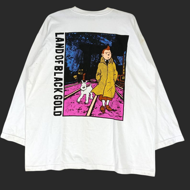 Puede incluir: Camiseta blanca de manga larga con un gr&aacute;fico del c&oacute;mic "Land of Black Gold". El gr&aacute;fico muestra un personaje con un abrigo amarillo con un perro blanco caminando sobre un camino rosa. El texto "LAND OF BLACK GOLD" est&aacute; impreso verticalmente.