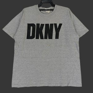 May include: A grey t-shirt featuring the black letters "DKNY" across the chest. The shirt has short sleeves and a crew neck. The t-shirt is hanging on a black hanger.