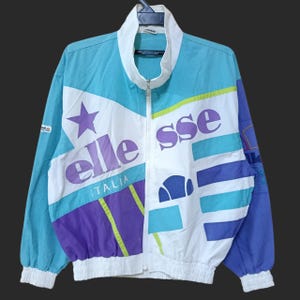 May include: A vintage ellesse Italia track jacket in a color block design. The jacket features a white, light blue, purple, and green color scheme with the brand name and star graphic. It has a full zipper and a stand-up collar.