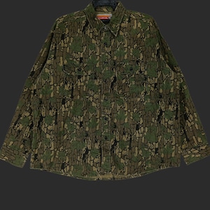 May include: A long-sleeved button-down shirt with a camouflage pattern featuring green leaves and brown bark-like textures. The shirt has two chest pockets and a collar. The overall design gives a woodsy, outdoorsy feel.