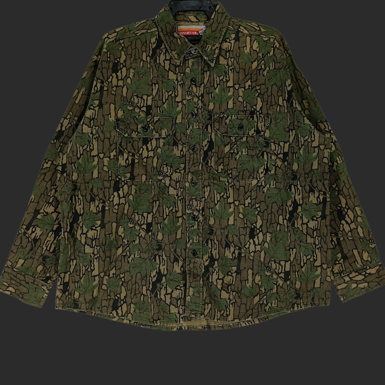 May include: A long-sleeved button-down shirt with a camouflage pattern featuring green leaves and brown bark-like textures. The shirt has two chest pockets and a collar. The overall design gives a woodsy, outdoorsy feel.