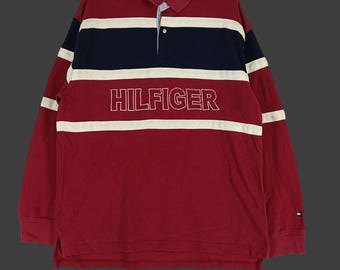 May include: A long-sleeved rugby shirt in red, navy blue, and white. The shirt features a collared neckline and the word "HILFIGER" in white block letters across the chest. White stripes accent the sleeves.