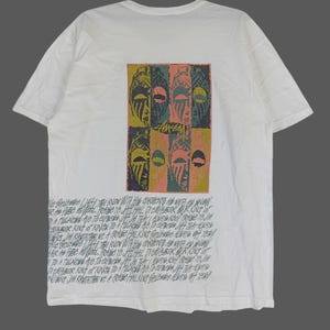 May include: White t-shirt featuring a graphic print of four stylised faces in black, green, yellow, and pink. The text "Amsan" is printed in black below the faces. The text "Le Gel JAH, YOU KNOW WITH HIS CARENTS AS WITH SNOW R A the Paseo PUBE NO WOOL Poise PRAISE YE JATI HALL DIE DIEMPEBOR OUR KING OF A TULNES AVe To express JIJI TRASLADUATION KOER YEJAM BOR KUNT OF KINGS TO A PULSO HE TO CARRER JAT THAT ALTI JAM RASTA TABI WE A PRAISE HALKING GLAY Off Te ALE BELAJAH YOU KNOW WITH WITH HIS GARMENTS AND WINE to S HAIR A PIBE NO WOOL PRAISE Ye ALH PRAISE YE HILFEL DEMPEROR GUR K DIEMPABOR KING OF KINGSIDA FI KINGST NUSING JAA RASTATABI WE A PRAS HALKING gelane ART off" is printed in black at the bottom of the shirt.