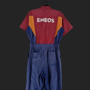 Może przedstawiać: Kombinezon z krótkim rękawem w kolorowym bloku. Góra jest bordowa z pomarańczowymi i niebieskimi akcentami, a dolna część jest niebieska. Słowo "ENEOS" jest wydrukowane na plecach na biało.