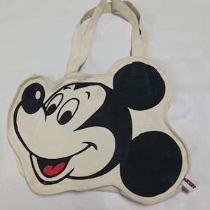 May include: A cream-colored canvas tote bag shaped like the head of Mickey Mouse. The bag features a large black and white graphic of the character's face with a red tongue. It includes two top handles and a small white tag with red text on the side.