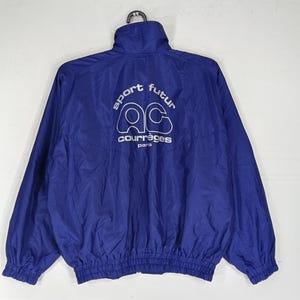 May include: A royal blue nylon jacket with a high collar and elastic cuffs. The back of the jacket features white text that reads "sport futur" above the "ac courr&egrave;ges paris" logo. The jacket is hanging from a black hanger.