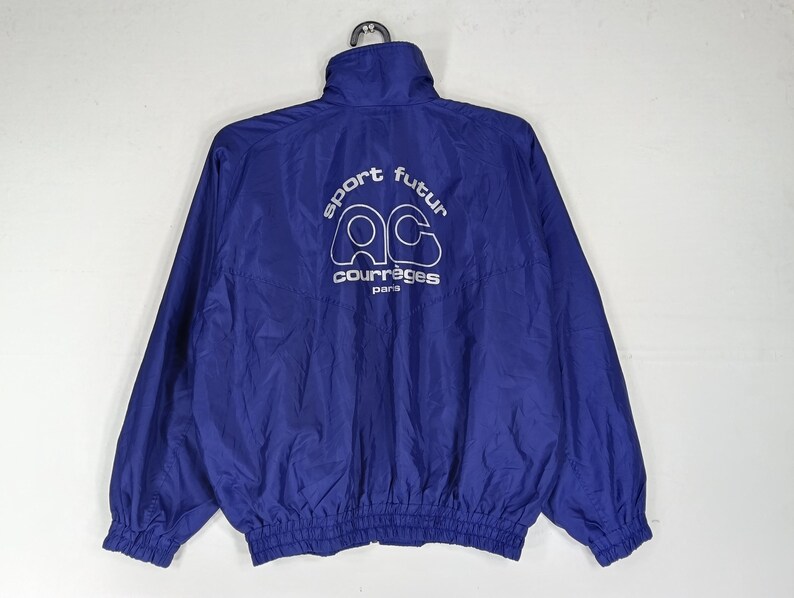 May include: A royal blue nylon jacket with a high collar and elastic cuffs. The back of the jacket features white text that reads "sport futur" above the "ac courr&egrave;ges paris" logo. The jacket is hanging from a black hanger.