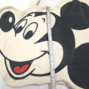 May include: A cream-colored tote bag featuring a large black and white graphic of Mickey Mouse with a red tongue. A measuring tape is placed vertically across the center, showing a length of approximately 12 inches from top to bottom edge.