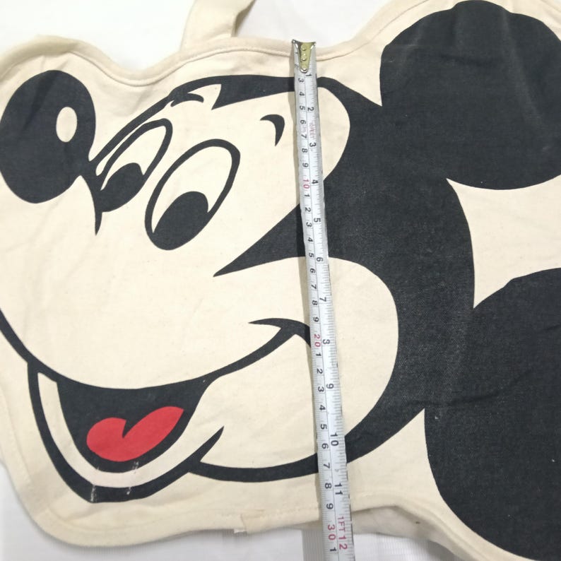 May include: A cream-colored tote bag featuring a large black and white graphic of Mickey Mouse with a red tongue. A measuring tape is placed vertically across the center, showing a length of approximately 12 inches from top to bottom edge.