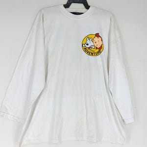 Puede incluir: Camiseta blanca de manga larga con un gr&aacute;fico circular que presenta un personaje de dibujos animados y un perro, con la palabra "ADVENTURE" en un c&iacute;rculo amarillo. La camiseta tiene cuello redondo y un corte holgado.