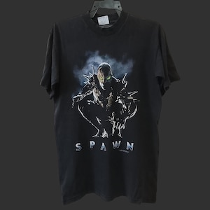 Puede incluir: Camiseta negra con un gráfico de Spawn. El diseño muestra al personaje en cuclillas sobre un fondo oscuro y ahumado. La palabra "SPAWN" está impresa en plata debajo. La camiseta está colgada en una percha negra.