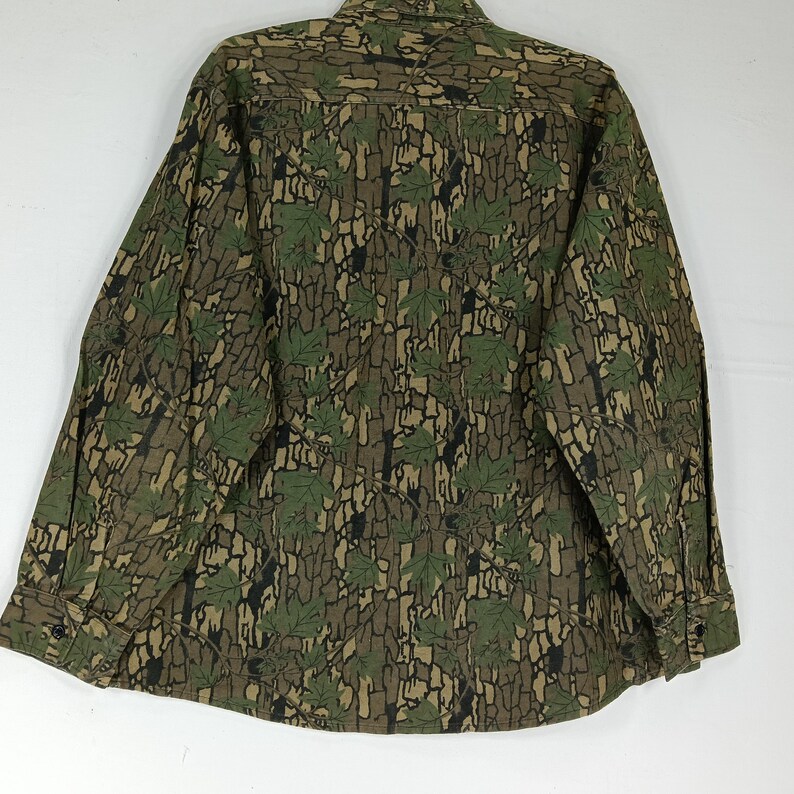 May include: A long-sleeved button-down shirt with a camouflage pattern. The shirt features a repeating design of green leaves and a bark-like texture in shades of brown and black. The shirt has a classic collar and buttoned cuffs.