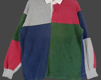 Può includere: Una maglia da rugby a blocchi di colore con colletto bianco e paramontura con bottoni. La maglia presenta pannelli grigi, rossi, verdi e blu. Le maniche lunghe hanno polsini grigi. La maglia è esposta su una gruccia nera.