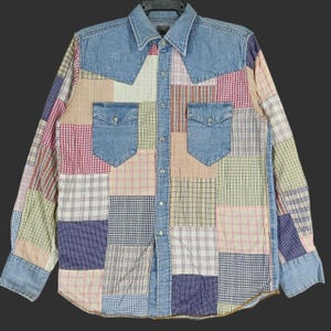 Könnte beinhalten: Ein langärmeliges Patchwork-Hemd mit Jeanskragen, Passe und Taschenklappen. Das Hemd weist eine Vielzahl von Stoffmustern in Blau-, Rot-, Rosa- und Beigetönen auf. Zwei Brusttaschen mit silbernen Knopfverschlüssen sind sichtbar.