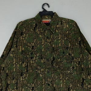 May include: A long-sleeved button-down shirt with a camouflage pattern featuring green leaves and brown bark-like textures. The shirt has two chest pockets and a collar. The brand name "Winchester" is visible on the collar.