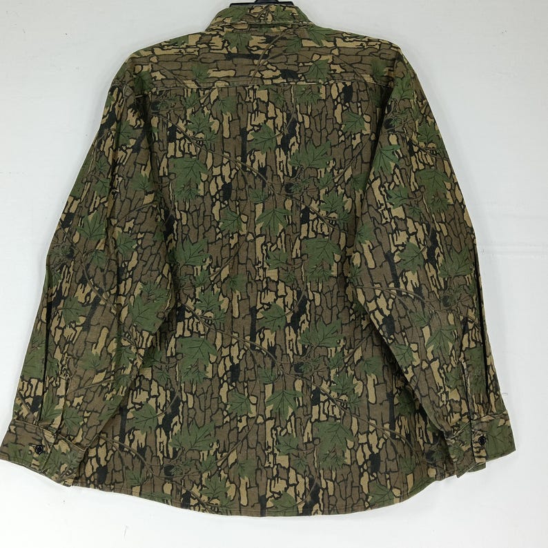 May include: A long-sleeved button-down shirt with a camouflage pattern. The shirt features a design of green leaves and brown bark-like textures. The shirt has a collar and buttoned cuffs. The overall color palette is earthy tones of green, brown, and black.