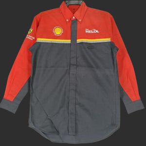 Puede incluir: Una camisa de trabajo de manga larga roja y gris, con el logotipo de Shell Helix y una franja amarilla en el pecho. La camisa tiene cuello abotonado y un pequeño bolsillo. El logotipo de Ferrari es visible en la manga izquierda.