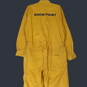 May include: A mustard yellow long-sleeve jumpsuit featuring a collar, a button-up front, and an elasticized waist. The back displays the text ROCK PAINT in bold black lettering. The garment includes two large back pockets and a small sleeve pocket.