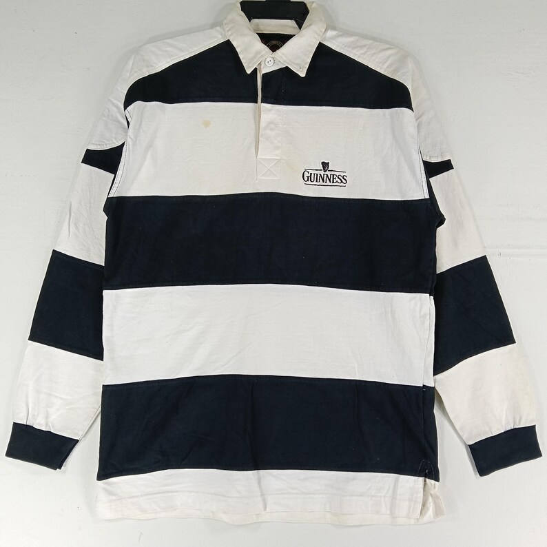 May include: A long-sleeved rugby shirt with alternating white and navy blue horizontal stripes. The shirt features a white collar and a three-button placket. The Guinness logo is embroidered on the chest.