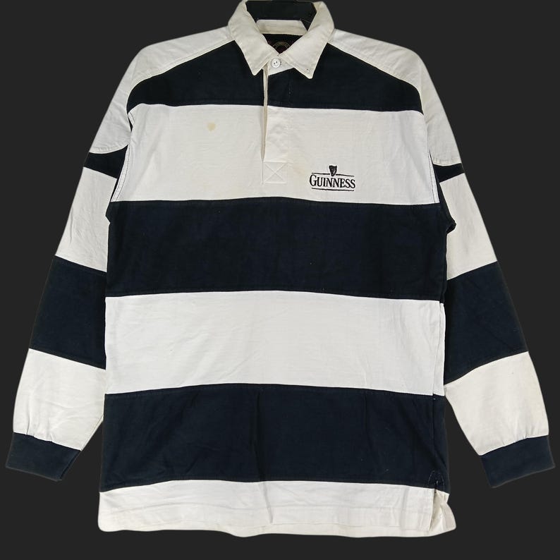 May include: A long-sleeved rugby shirt with alternating black and white horizontal stripes. The shirt features a collared neckline, a three-button placket, and the Guinness logo embroidered on the chest.