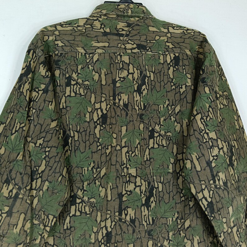 May include: A button-down shirt with a camouflage pattern featuring brown, green, and black tones. The design incorporates a bark-like texture with green leaf accents, creating a nature-inspired aesthetic. The shirt has a classic collar and long sleeves.