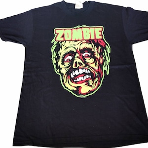 Puede incluir: Camiseta negra con un gráfico verde, rojo y amarillo de una cara de zombie y la palabra "ZOMBIE" en letras verdes.