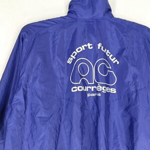 May include: A royal blue jacket with a high collar. The back of the jacket features the white text "sport futur" above the white logo "AC" and the word "courr&egrave;ges paris". The jacket is made of a lightweight, possibly nylon, material.