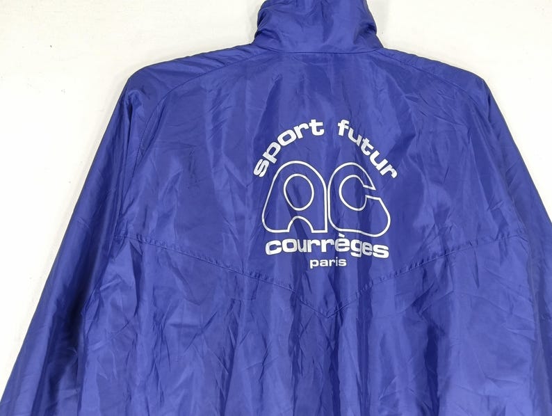 May include: A royal blue jacket with a high collar. The back of the jacket features the white text "sport futur" above the white logo "AC" and the word "courr&egrave;ges paris". The jacket is made of a lightweight, possibly nylon, material.