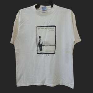 May include: Off-white t-shirt with a black and white graphic of a person in a landscape. The graphic reads "JACKSON BROWNE I'm Alive" and "SUMMER '94". The shirt has short sleeves and a round neck.