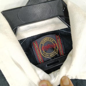 May include: Close-up of a black and white collared shirt with a Guinness label. The label is rectangular with a gold border and features the Guinness logo in red and gold. The shirt is hanging on a black hanger.