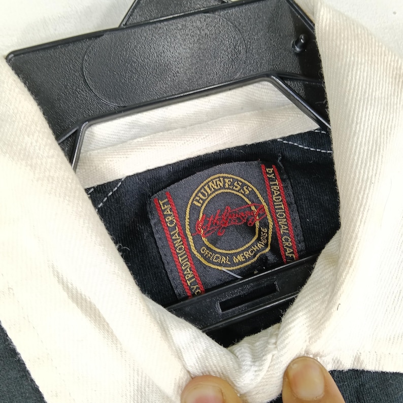 May include: Close-up of a black and white collared shirt with a Guinness label. The label is rectangular with a gold border and features the Guinness logo in red and gold. The shirt is hanging on a black hanger.