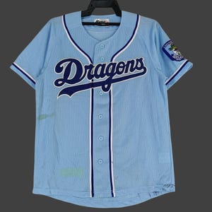 Puede incluir: Camiseta de béisbol azul claro con la palabra "Dragons" en escritura azul marino en el pecho. La camiseta tiene ribetes blancos y azul marino, una parte delantera abotonada y un parche con el logotipo del equipo en la manga.
