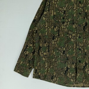 May include: A long-sleeved button-down shirt with a camouflage pattern. The shirt features a mix of brown, green, and black colors, with a leaf design. It has a chest pocket and black buttons.